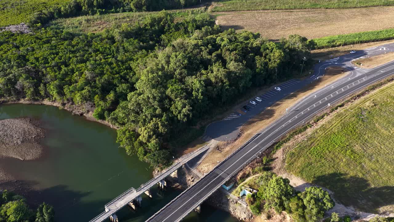 Drone footage captures a bridge over a river surrounded by dense greenery and fields in Port Douglas, Australia