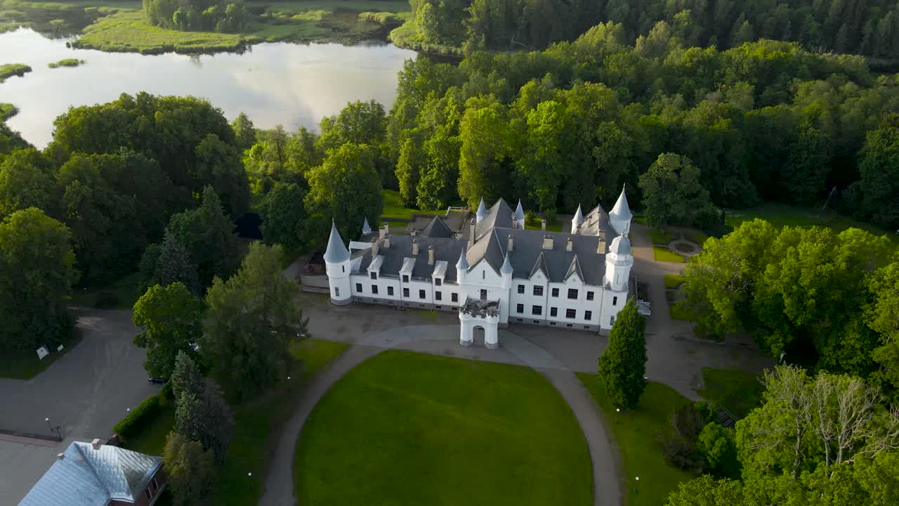 Drone orbit view over scenic Alatskivi historic castle on the hill, Estonia. Forested white manor among green trees and glowing lake at sunset. Rural tranquil landscape, poetic charm of grand estate