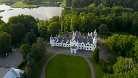 drone orbit view over scenic alatskivi historic castle on the hill, estonia. forested white manor among green trees and glowing lake at sunset. rural tranquil landscape, poetic charm of grand estate