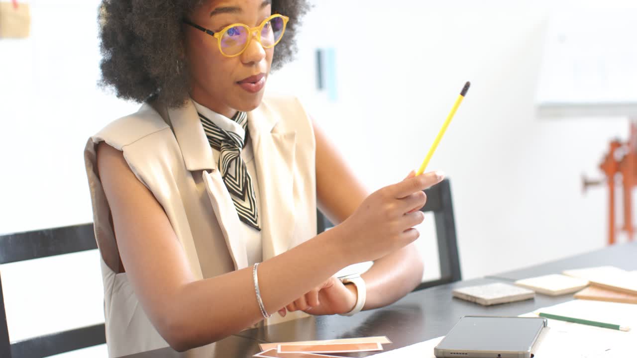 Donna afroamericana con gilet e sciarpa in studio che legge campioni su tablet, facendo roteare una matita e scegliendo le tonalità.