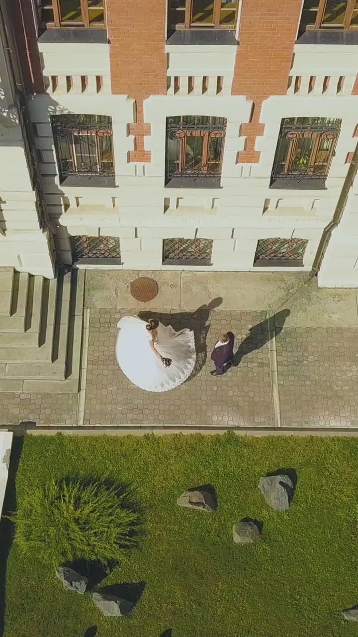 distant bride in white fluffy dress spins near groom at modern building facade and green lawn on sunny day upper view