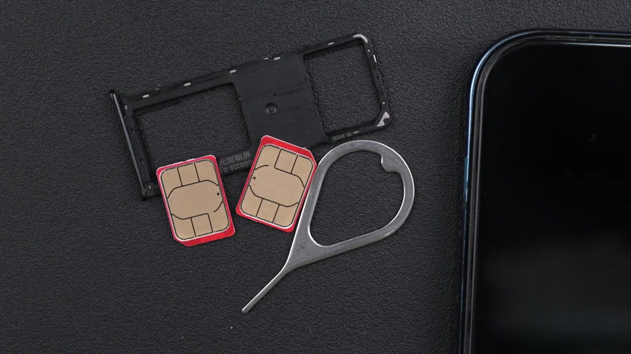 Dois cartões SIM com bordas vermelhas, uma bandeja de SIM e uma ferramenta ejetora estão dispostos ao lado de um smartphone em uma superfície preta texturizada. Útil para conteúdo focado em tecnologia
