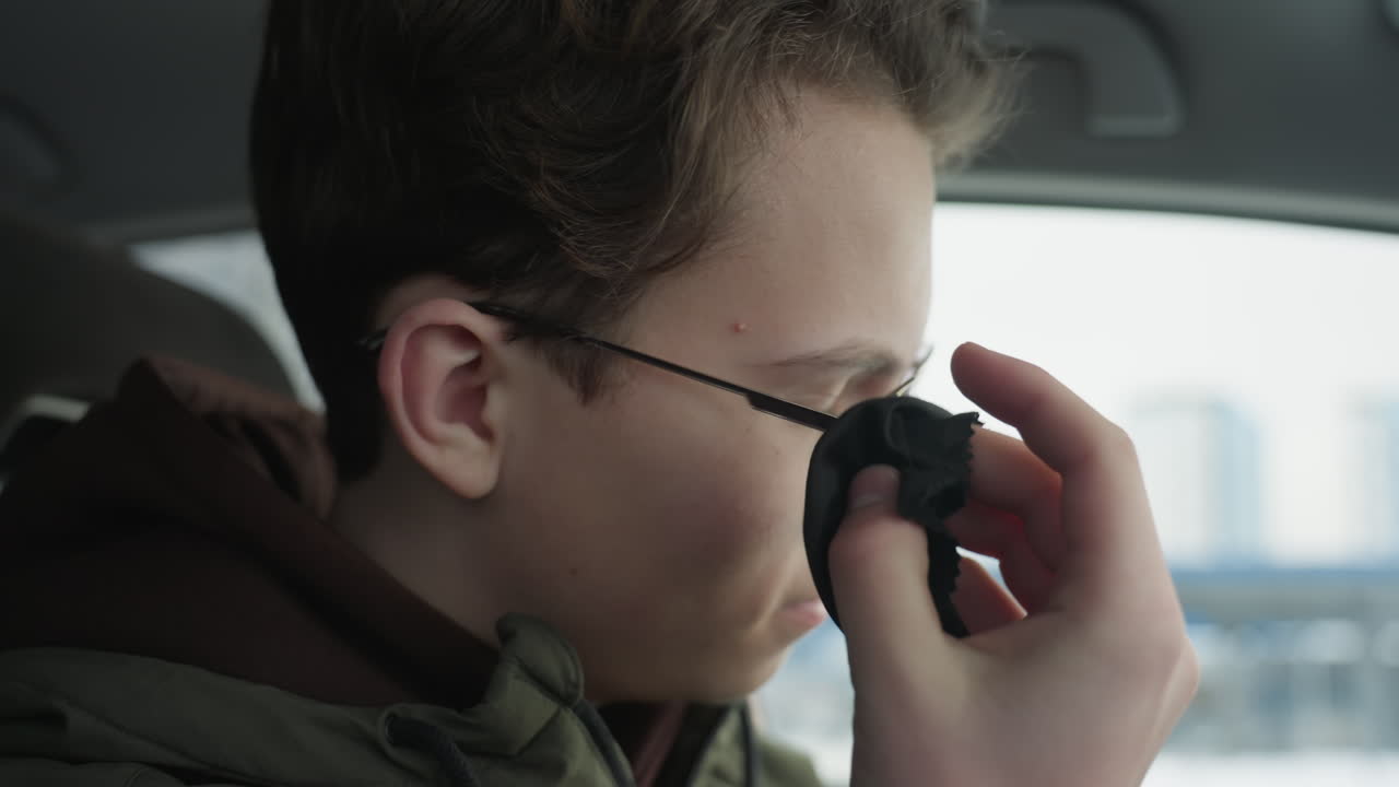 Young man cleaning glasses lens with microfiber cloth while seated in car interior, blurred building visible through window providing context for eyewear maintenance under soft natural light