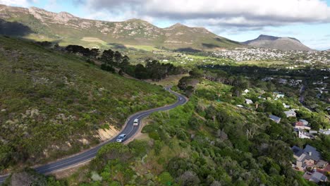 Toma-Aérea-Inversa-De-Camiones-Y-Autobuses-Conduciendo-Por-Una-Carretera-De-Montaña-Con-Curvas-Sobre-Una-Ciudad-Con-Nubes-Y-Sombras,-Cerca-De-Ciudad-Del-Cabo-Sudáfrica