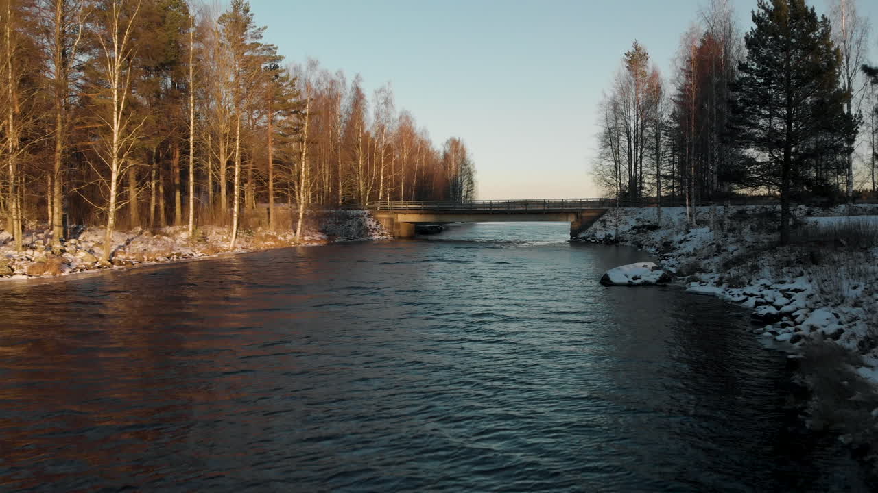 Ilmakuva dronella, joen yllä, kohti siltaa, jota ympäröi lehdettömän metsän ja ensilumen peittämä maa, aurinkoisena talvipäivänä, lähellä Joensuuta, Pohjois-Karjalassa, Suomessa