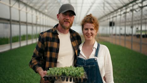 portrait of a happy farmer guy in a cap with a beard and a woman with red hair posing in a greenhouse on a farm among plant sprouts