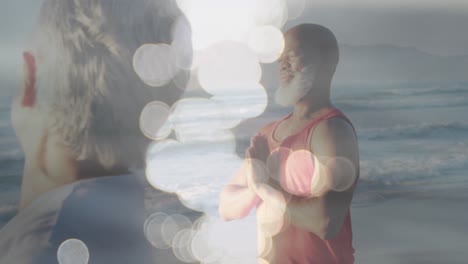 animation of glowing spots over happy african american senior couple practicing yoga on beach