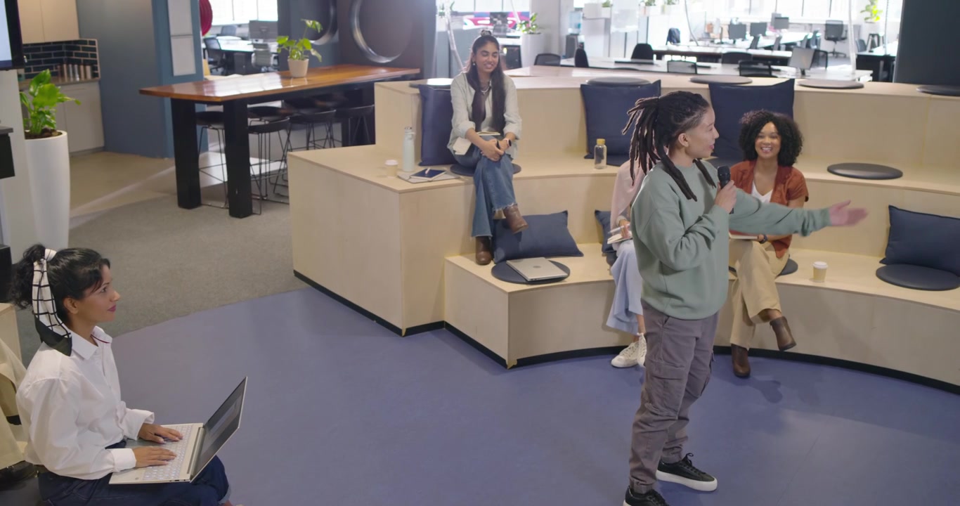 Diverse coworkers clapping at office, nonbinary presenter rising, walking center, speaking into mic