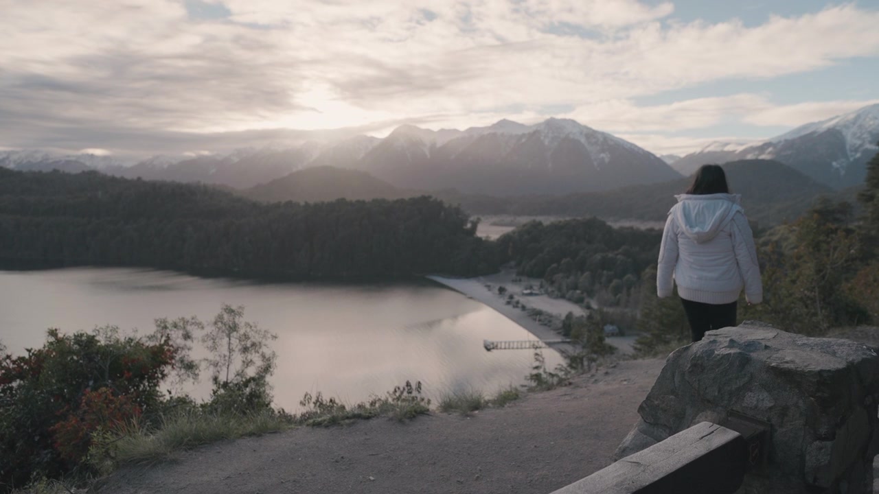 A woman in a white jacket walks along a viewpoint overlooking Lake Espejo in Villa la Angostura, Argentina. The scene captures the serene beauty of the landscape, ideal for travel content.