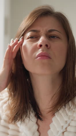 mujer en túnica mira la cara en el espejo después de ir al centro de spa. atractiva dama con cabello oscuro mira el efecto después de la máscara facial en el área de spa espaciosa