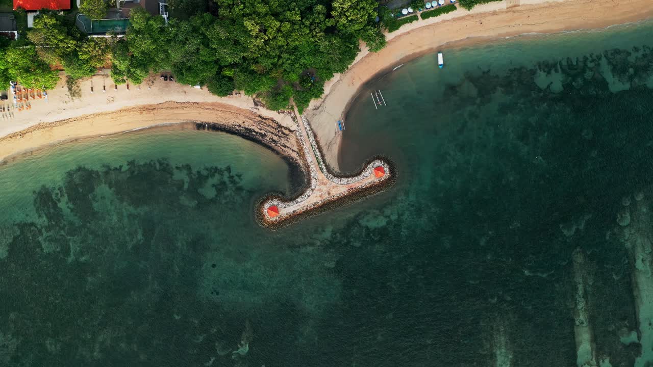Aerial drone view of Bali tropical shoreline featuring twin gazebos built on a stone breakwater, calm turquoise waters, golden sandy coves, and lush green canopy blending into coastal charm