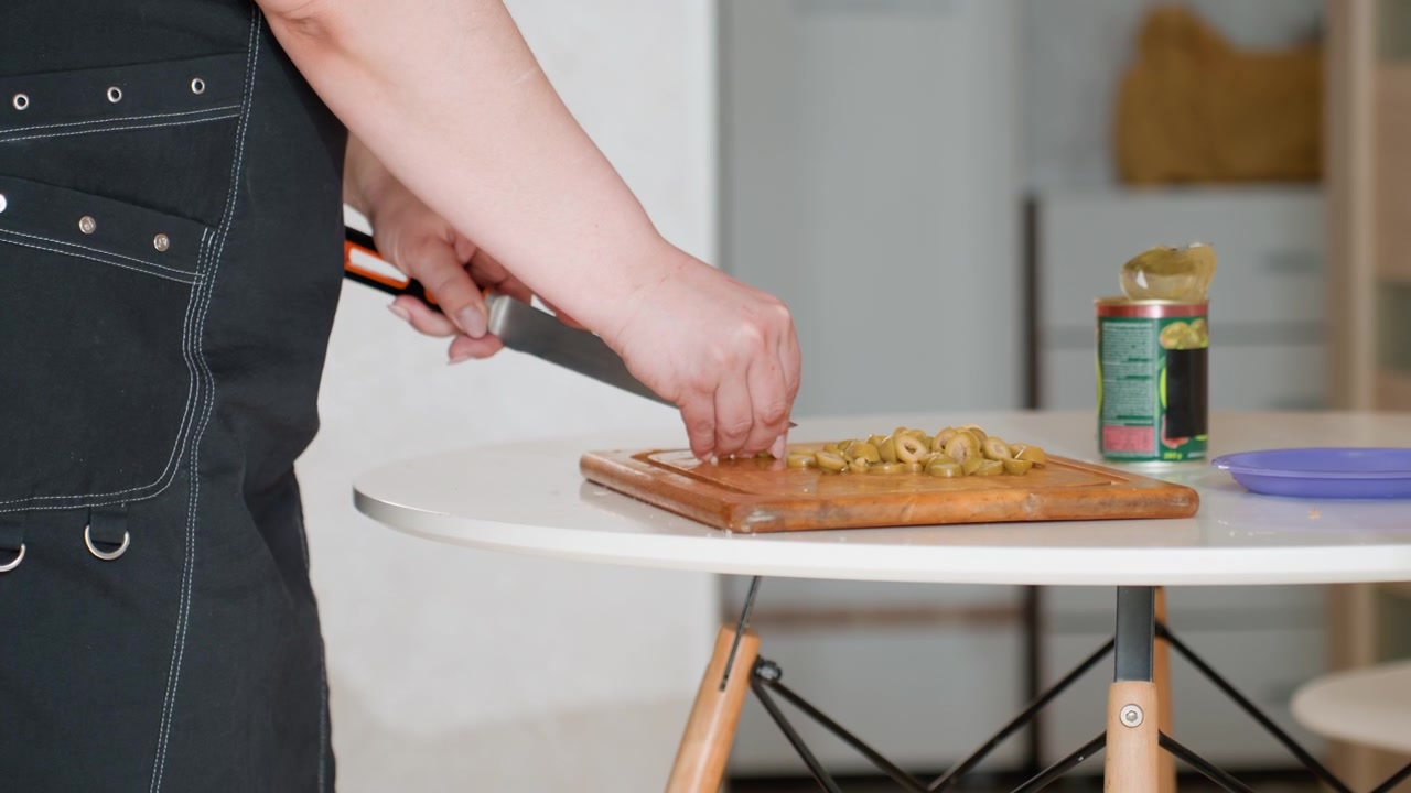 Tampilan dekat tangan berapron gelap sedang menyelesaikan topping pada roti pipih atau pizza tipis di meja bundar di dapur rumah, dengan gerakan mencubit, menabur, menekan, dan meratakan dalam cahaya siang yang lembut.