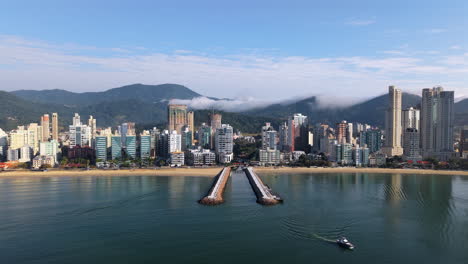 slow drone pans itapema meia praia skyline and beachfront in paraná, brazil, showcasing sandy beach, oceanfront high-rises, marina piers, and the serra do mar mountains under a partly cloudy blue sky