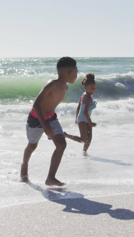 hermanos afroamericanos sonrientes corriendo y usando trajes de baño en una playa soleada