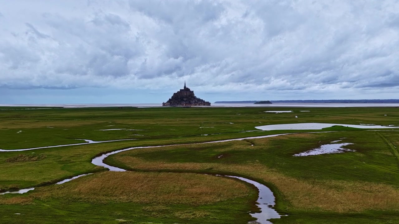 Vidvinkeludsigt over en fjerntliggende ø og græsklædt kystland under en overskyet himmel.