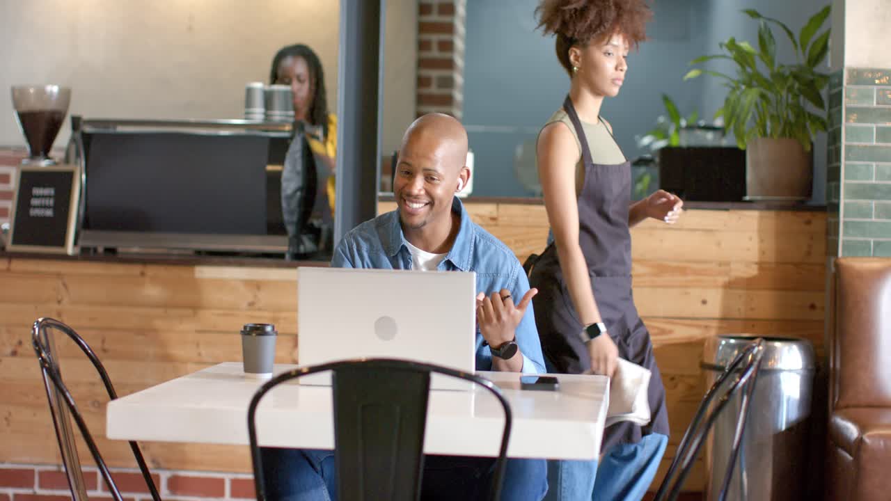 African American man waving on call, barista crossing behind chair, typing on silver laptop at cafe