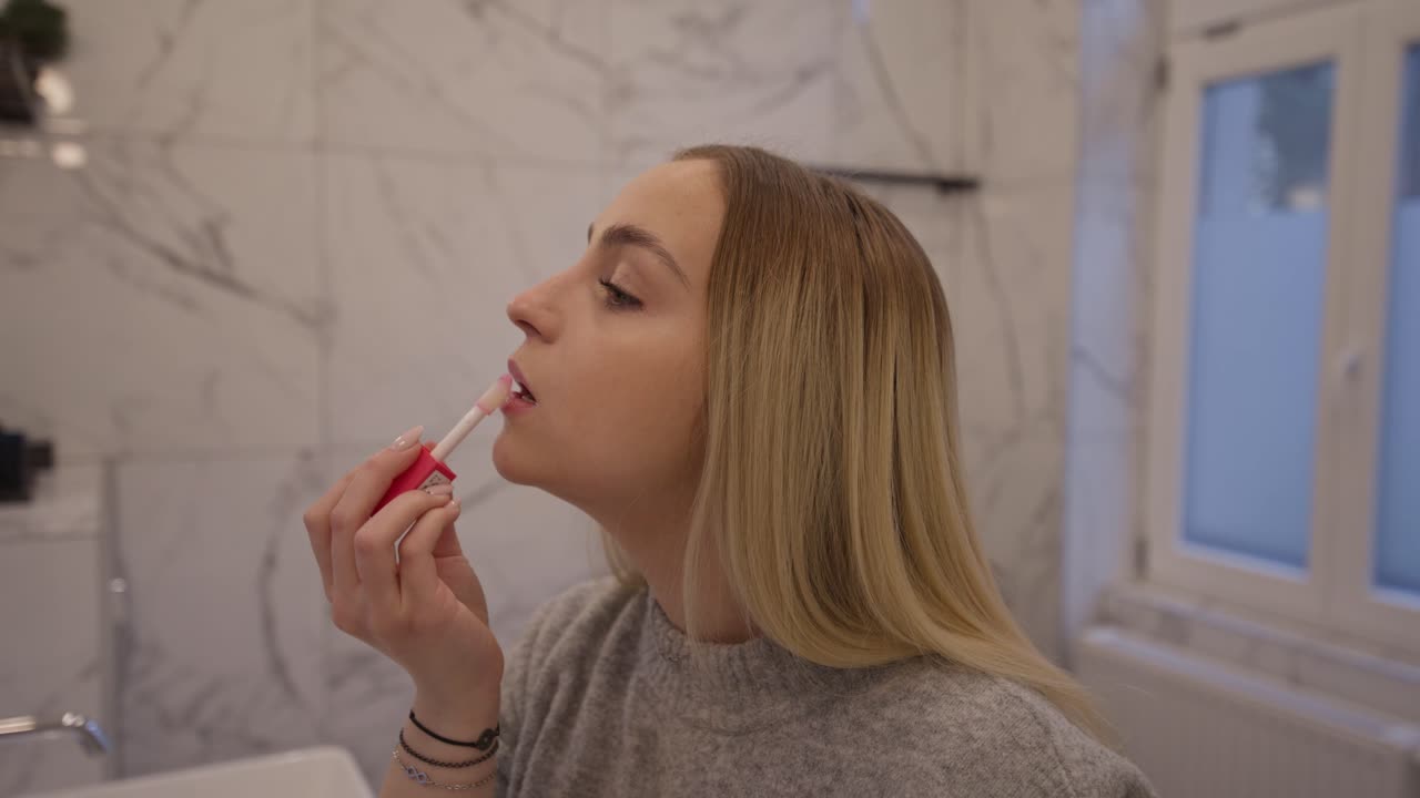 A young blonde woman applies lip gloss while standing in a modern bathroom. She looks at her reflection with focus, highlighting a daily beauty and skincare routine at home