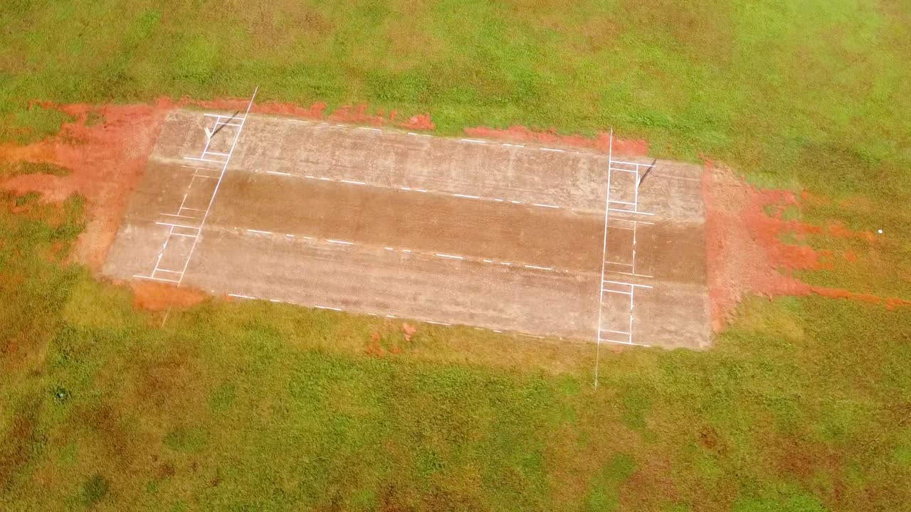 Ripresa aerea con drone che ruota su un campo da cricket rustico, segnato a mano, in un campo verde di campagna con dettagli di terra rossa.