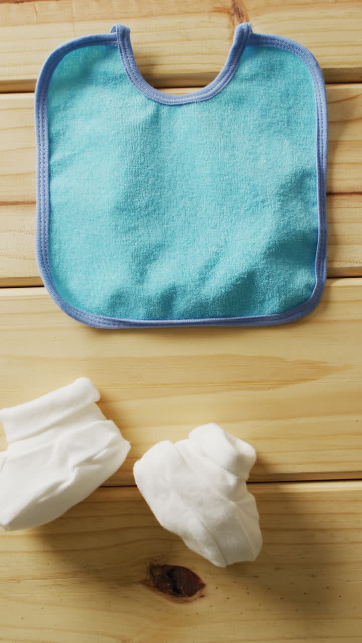 Vertical video of close up of white booties and blue bib on wooden background with copy space