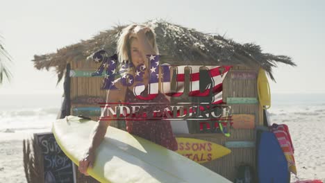 Bandera-Americana-Ondeando-Sobre-El-Texto-Del-Día-De-La-Independencia-Contra-Una-Mujer-Caucásica-Con-Tabla-De-Surf-En-La-Playa