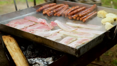 tocino y salchichas fritos en una sartén a fuego abierto - desayuno de camping