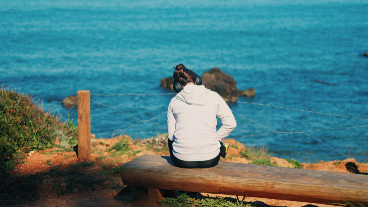 Woman with headphones on her head sitting on a wooden bench with a view of the sea