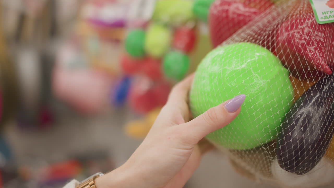Hand view of female manicured hands gently hitting rubber toy ball in mesh packaging, exploring tactile texture and round shape in retail store with blurred colorful displays under ambient lighting
