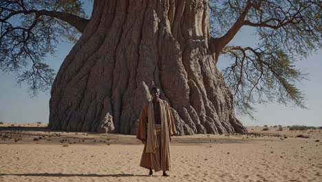 wide shot of a man standing in the desert under the shade of a giant baobab tree, showcasing the stark beauty and resilience of nature in arid environments