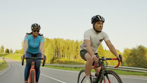 handsome bearded professional male cyclist riding his racing bicycle in the morning together with his girlfriend both wearing protective helmets and eyeglasses sun shining through between them