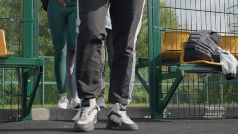 lower angle view capturing feet in sneakers as people in joggers walk into sports court, detailed with green gate and chairs with bags, ready for activity