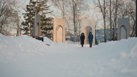 snowy winter day with two people in hooded jackets walking side by side on snow covered path near tall concrete monument structures, surrounded by bare trees under soft evening light
