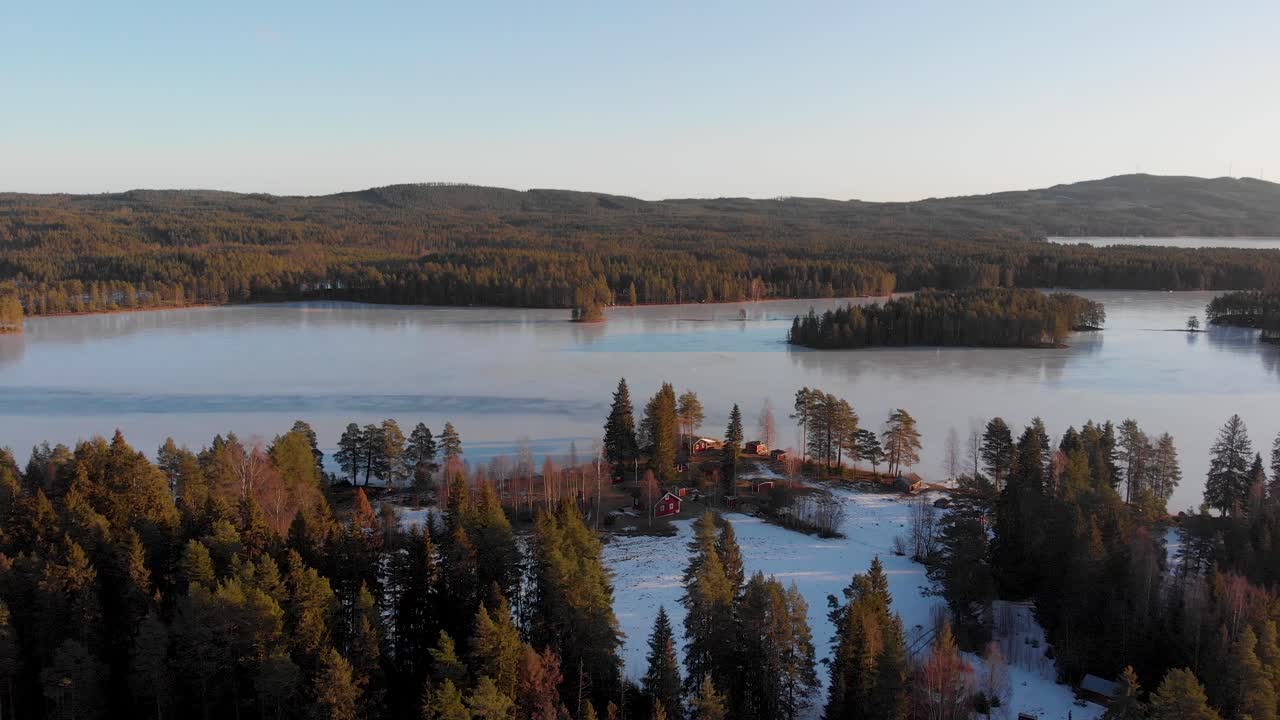 Langsames Aufsteigen an einem klaren Wintertag mit blauem Himmel, eisbedecktem See im Hintergrund und einer Gruppe roter Häuser in Appelbo, Vansbro Kommun.