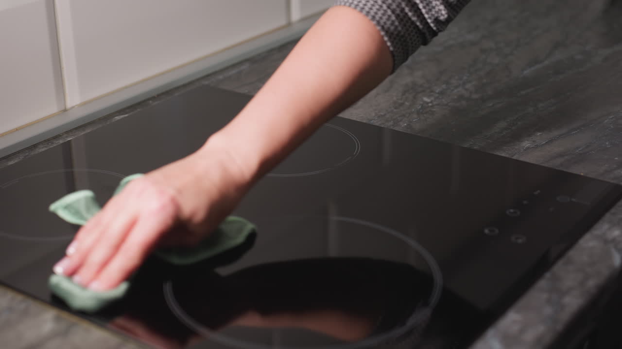 Person cleaning electric stove glass surface with green cloth in modern kitchen showing hand movement across stovetop maintaining hygiene