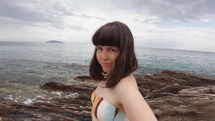 Una giovane donna bruna con la frangia che sorride e si diverte davanti alla telecamera su una spiaggia rocciosa in Grecia in una giornata di sole