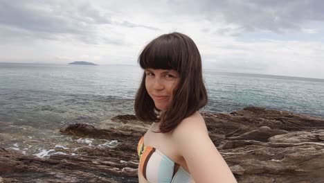 a young brunette woman with bangs smiling and having fun at the camera on a rocky beach in greece on a sunny day