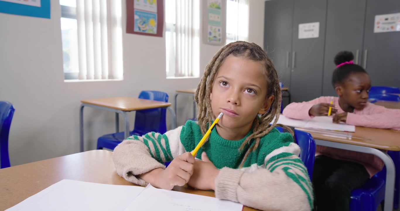 Ragazzo afroamericano in primo piano che lavora in classe su un foglio, segnando e riflettendo con la matita, con i compagni di classe sullo sfondo.
