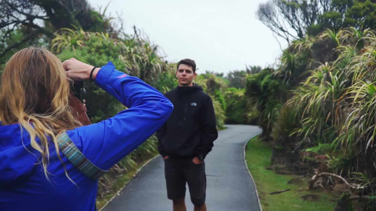 Young caucasian female from behind takes photo of male tourist at Punakaiki Pancake Rocks, New Zealand