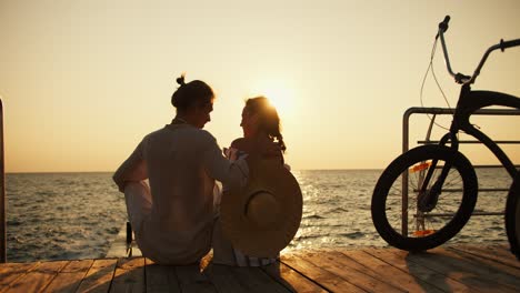 Herauszoomen:-Mann-Und-Mädchen-Sitzen-Auf-Dem-Pier-Am-Strand-Gegenüber-Dem-Sonnenaufgang-Und-Dem-Meer.-Ein-Glückliches-Paar,-Ein-Mann-In-Leichter-Kleidung-Und-Ein-Mädchen-Mit-Strohhut,-Sind-Bei-Sonnenaufgang-Mit-Fahrrädern-Angekommen-Und-Sitzen-In-Der-Nähe-Des-Meeres