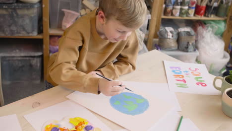 Niño-Rubio-Pintando-Una-Tierra-En-Un-Papel-Sentado-En-Una-Mesa-En-Un-Taller-De-Artesanía-Donde-Hay-Carteles-Con-Citas-Ambientales-1