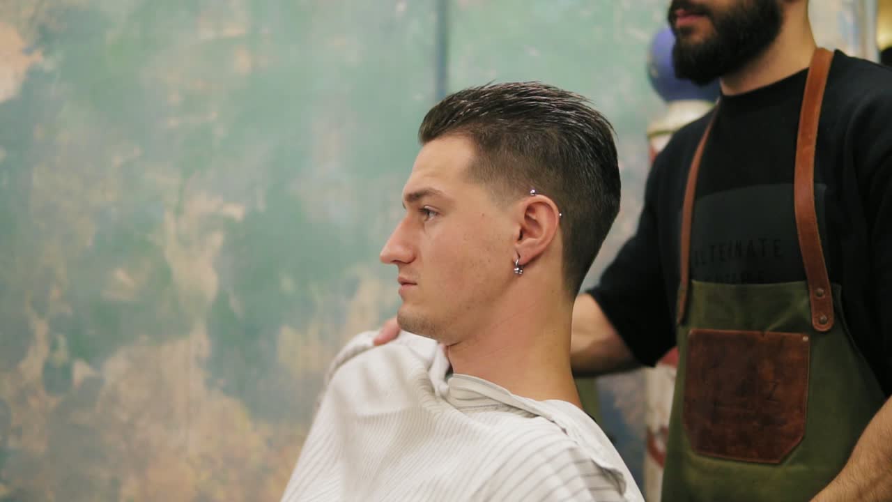 joven barbero barbudo quitando la capa del salón del elegante cliente con perforación en la oreja y tatuaje. cliente satisfecho