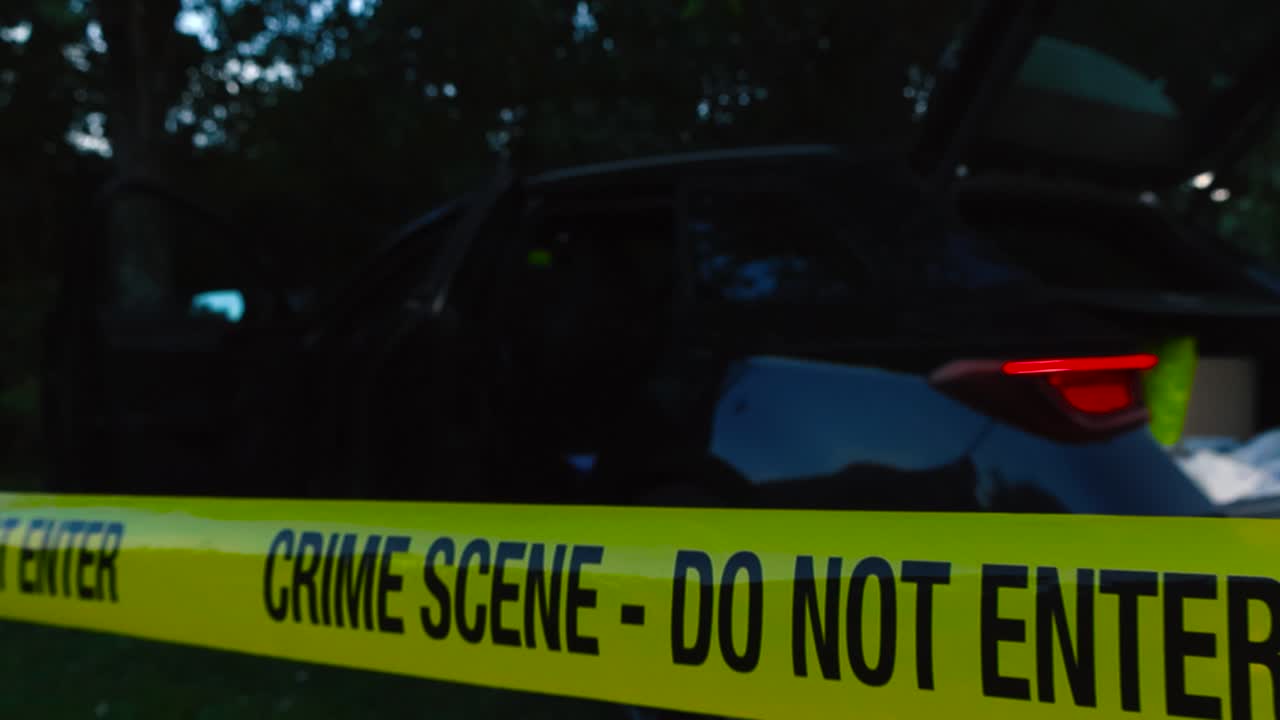 Close up view of a yellow colored police investigation forensics Crime Scene tape pulled in front of a crashed black vehicle during night time, cars indicator lights are blinking and flashing, trees