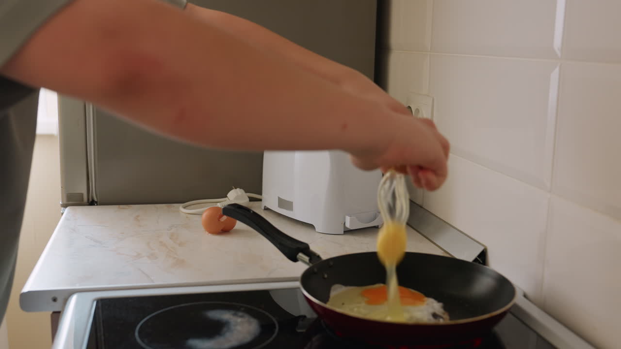 Hand view of kid breaking egg into frying pan on hot stove in bright home kitchen with toaster on counter and sunlight shining through window creating warm morning cooking mood