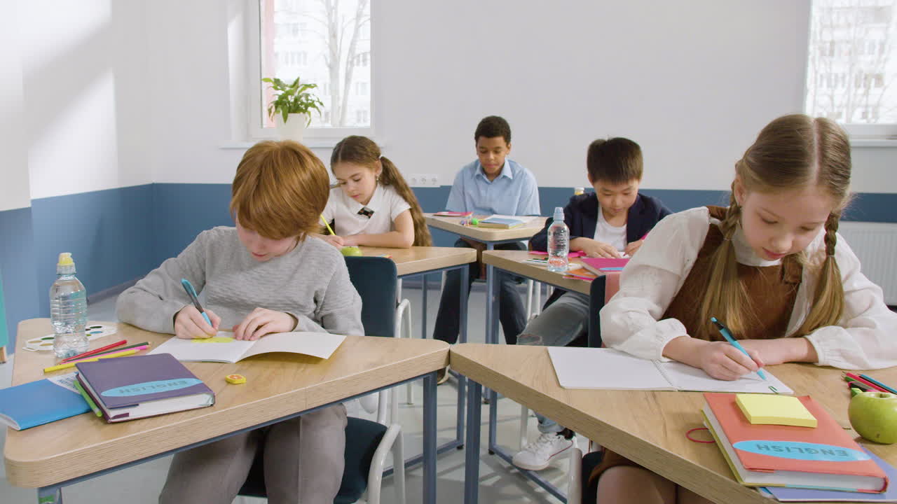 Multiethnic Group Of Students Sitting At Desks In English Classroom Raising Their Arms To Answer The Teacher's Question