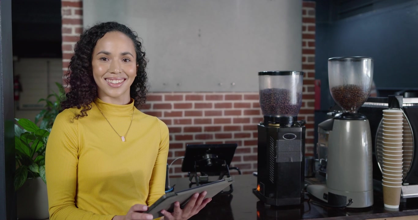Donna afroamericana con un dolcevita color senape che controlla un tablet, elaborando ordini vicino ai macinacaffè del bar