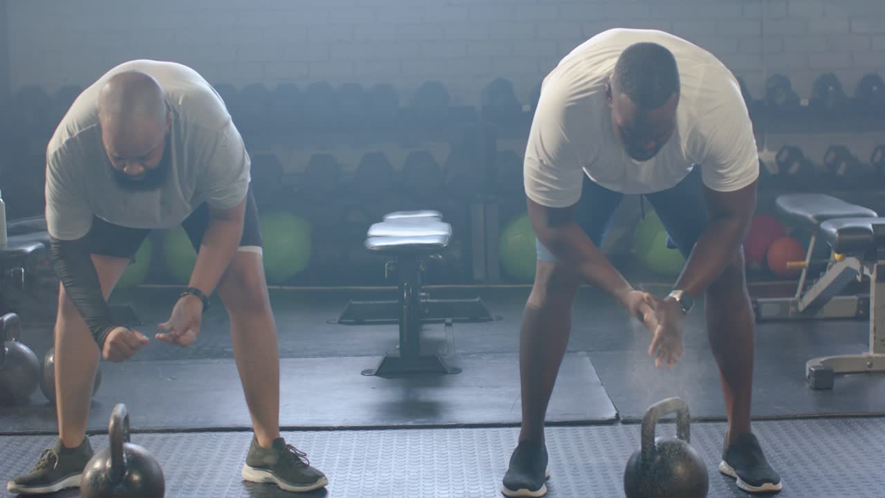 Two men clapping, creating chalk dust, hinging and reaching, gripping kettlebells at gym for swing