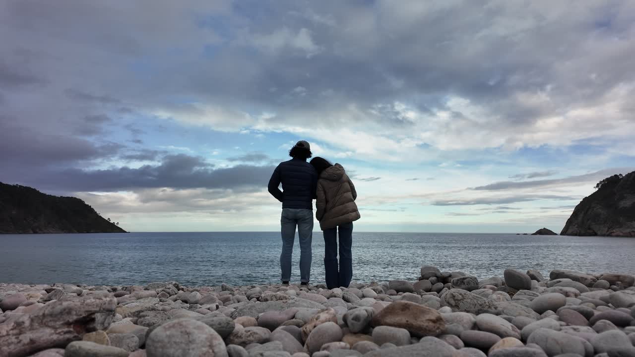 A couple stands on a rocky shoreline facing the sea under a dramatic cloudy sky