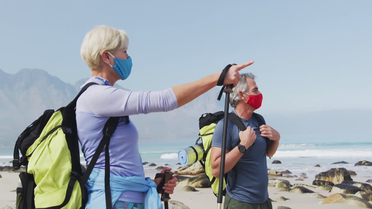 Coppia di escursionisti anziani con mascherine, zaini e bastoni da trekking che indicano una direzione.