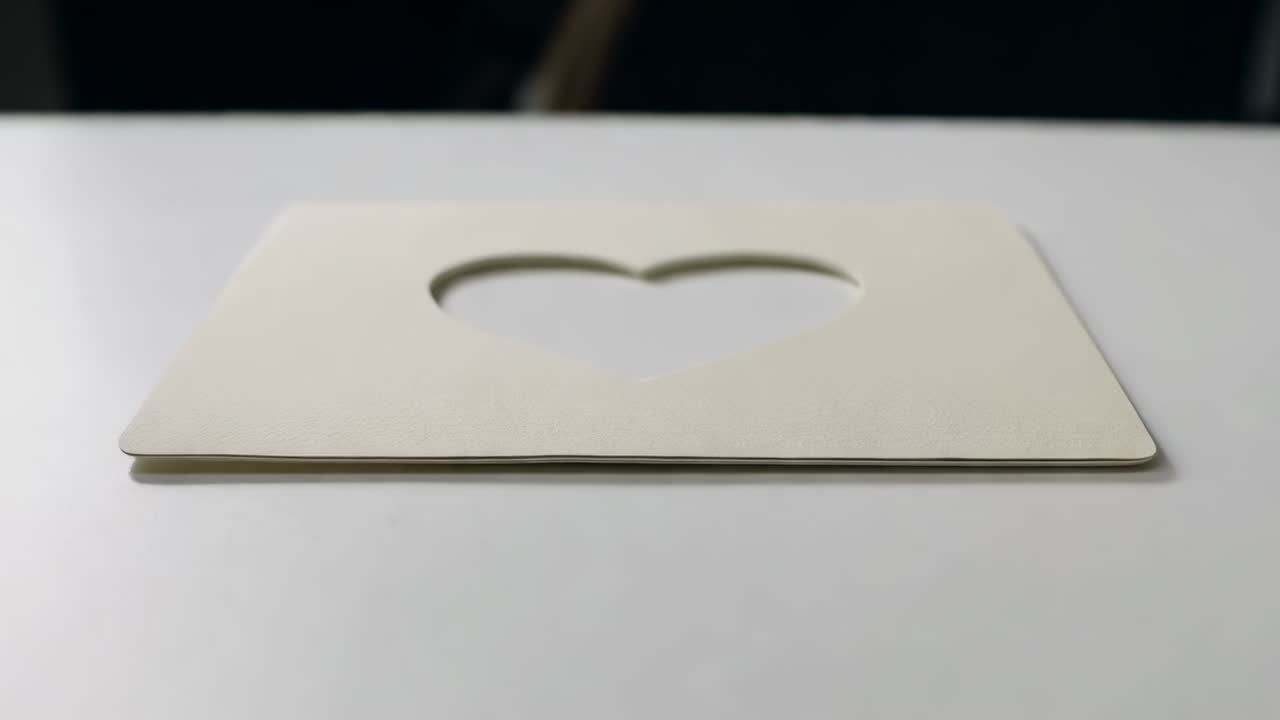 Telecamera in movimento che si avvicina a un biglietto color crema su un tavolo bianco, inquadrando un cuore ritagliato, ombra interna, banda scura