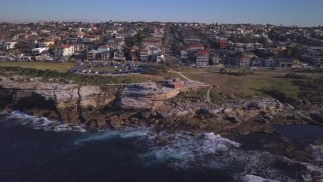 Flying-sideways-aerial-shot-of-ocean-waves-crashing-at-seashore-and-rockpool-near-waterfront-houses-and-beach