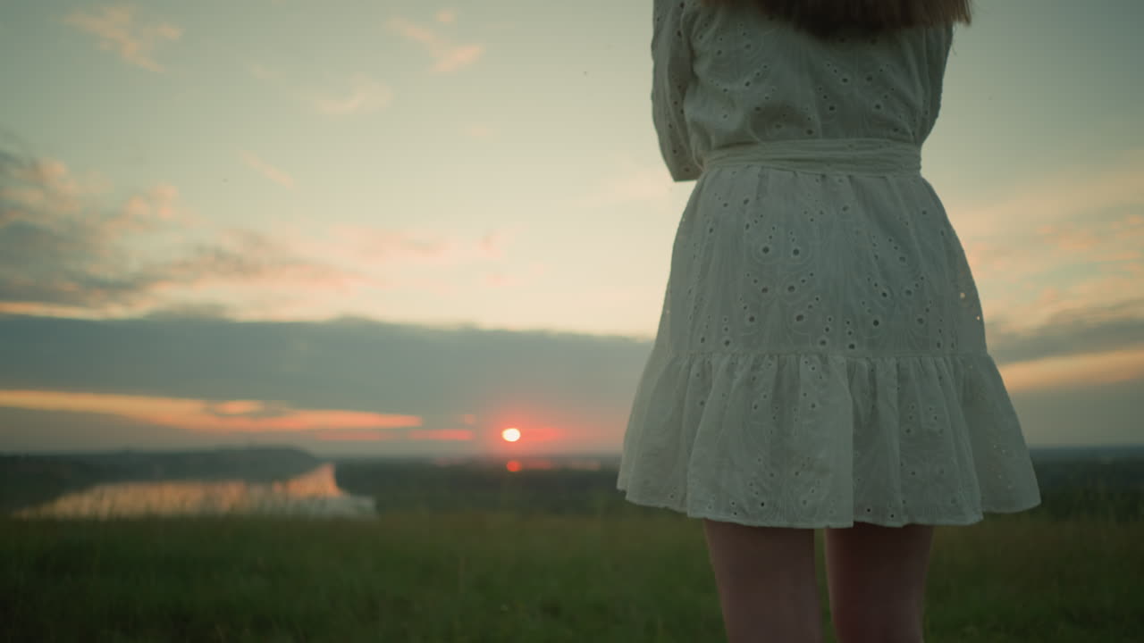 Low-angle shot on the lower body of a woman in a white dress standing alone in a serene grassy field, arms folded, as she gazes over a tranquil lake during a peaceful sunset calmness and contemplation